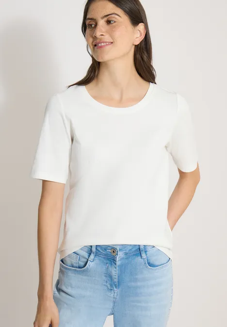 Frau trägt weißes T-Shirt und blaue Jeans vor hellem Hintergrund, Hand in der Tasche.