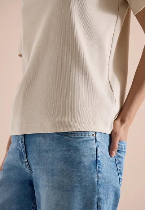 Close-up van een persoon in een beige t-shirt en blauwe denim short, hand in zak, neutrale achtergrond.