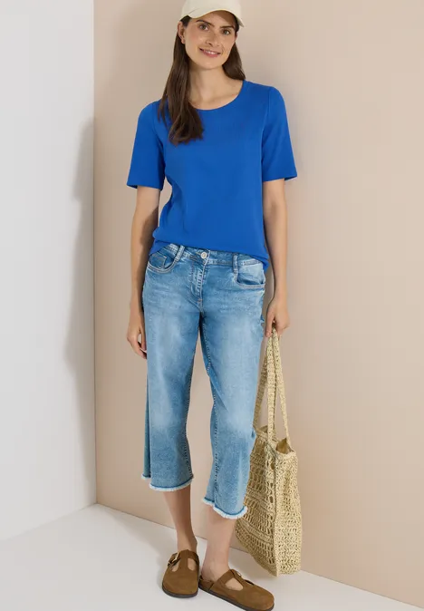 Basic Shirt met geribde textuur surf blue Vrouw in blauw T-shirt, caprijspantalon, strooien tas en bruine sloffen voor een neutrale muur.