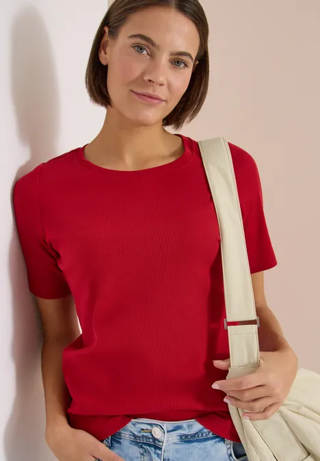 Person lehnt an Wand, trägt rotes T-Shirt, blaue Jeans und eine helle Schultertasche.