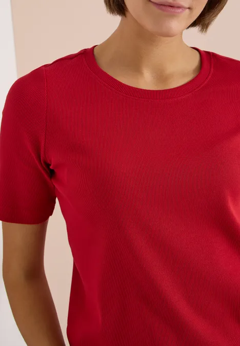Frau trägt rotes T-Shirt mit Rundhalsausschnitt, kurzärmlig, vor hellem Hintergrund.