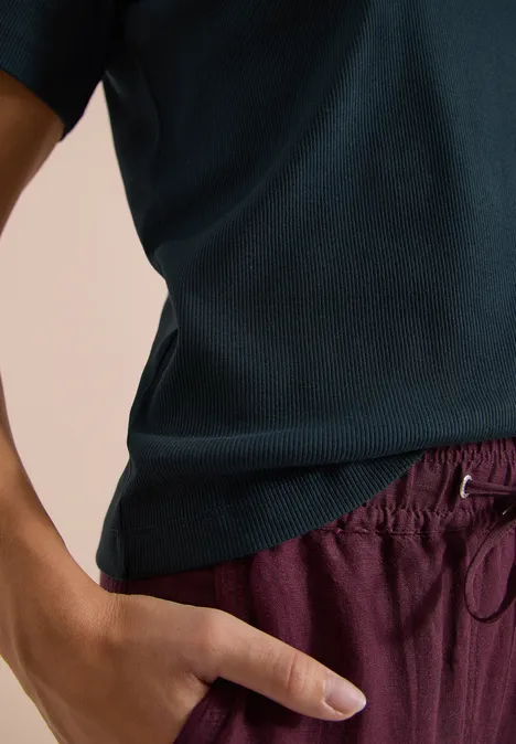 Person trägt ein dunkelblaues Shirt und bordeauxrote Hose, Hand in Tasche.