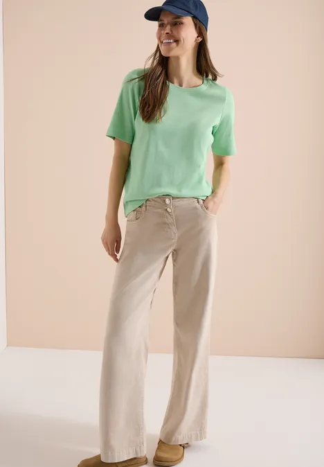 Basic geribd Shirt juicy lime Vrouw in groen T-shirt, brede beige broek, witte sneakers en blauwe pet voor een neutrale achtergrond.