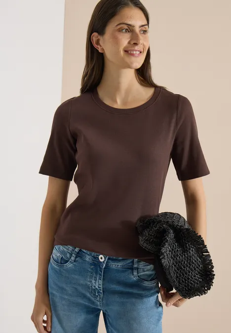 Eine schlanke Frau in braunem T-Shirt und blauen Jeans, die eine schwarze gehäkelte Tasche hält.