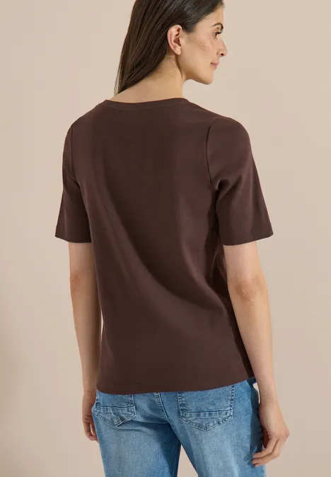 Frau steht mit dem Rücken zur Kamera, trägt braunes T-Shirt und blaue Jeans vor hellem Hintergrund.