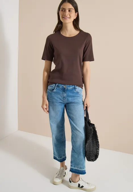 Frau in braunem T-Shirt und blauen Jeans, weiße Sneaker, schwarze Tasche, stehend vor hellem Hintergrund.