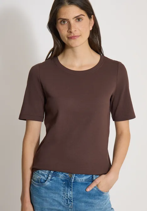 Frau trägt braunes Kurzarmshirt und blaue Jeans, eine Hand in der Tasche.