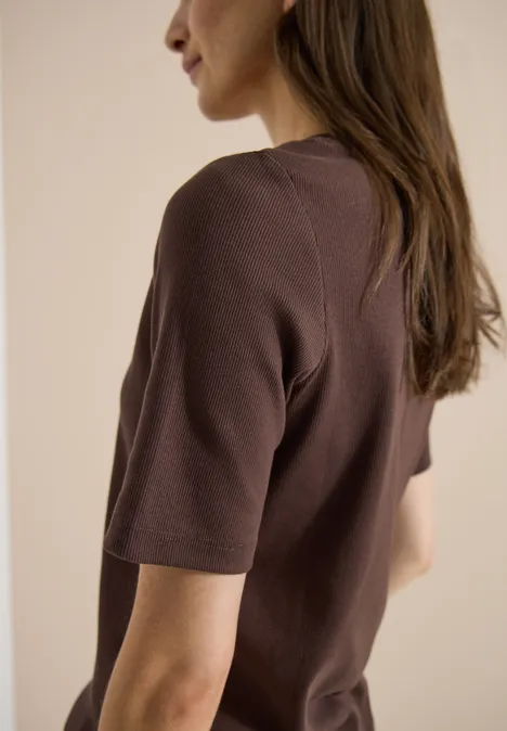 Eine Frau von hinten mit braunem T-Shirt, lange dunkle Haare, neutrale Wand im Hintergrund.