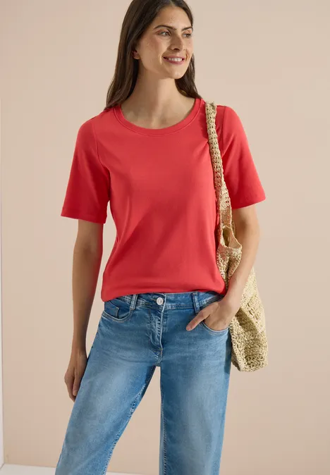 Frau in rotem T-Shirt und blauen Jeans mit Strohtasche über der Schulter, lächelt.