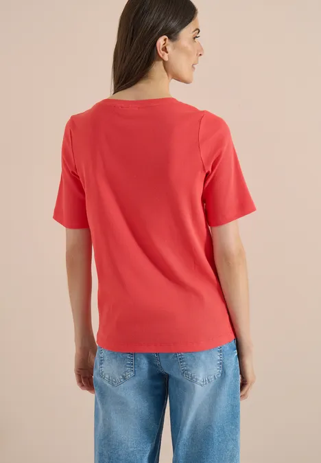 Eine Person von hinten im roten T-Shirt und blauen Jeans vor neutralem Hintergrund.