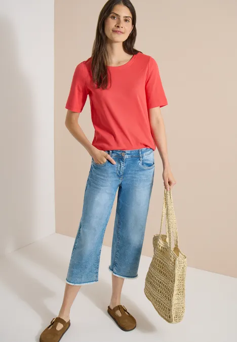 Frau in rotem Shirt, weitem Jeans-Culotte, braunen Schuhen und Korbtasche.