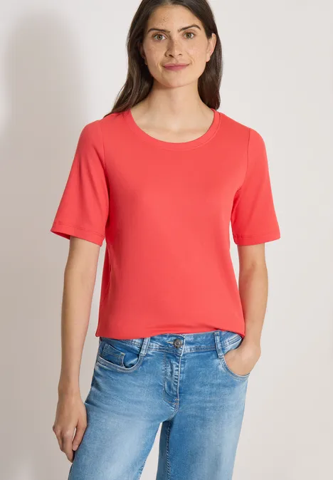 Frau in rotem T-Shirt und blauer Jeans vor neutralem Hintergrund.