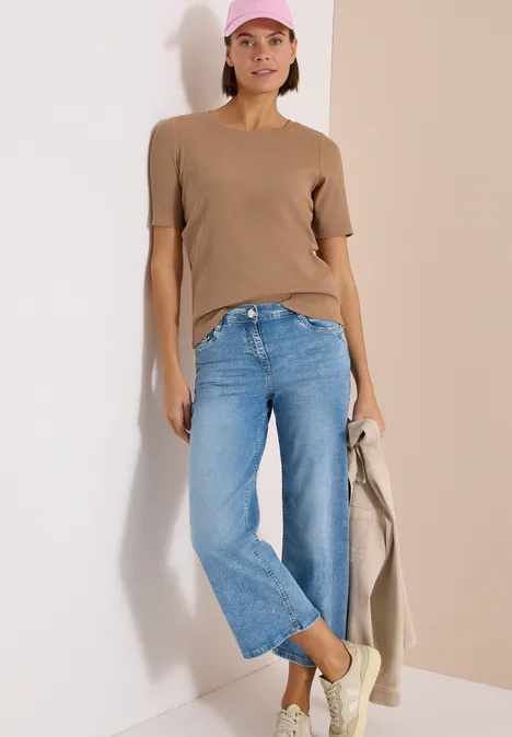 Persoon leunt tegen een lichte muur, draagt bruin T-shirt, blauwe jeans en houdt een beige jas vast.
