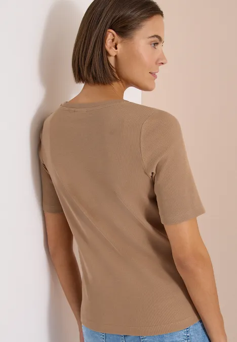 Vrouw van achteren met korte bob, draagt een beige shirt met korte mouwen en leunt tegen een witte muur.