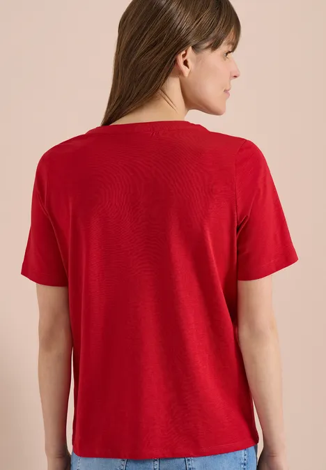 Frau von hinten in rotem Kurzarmshirt, Jeans und weißen Sneakern vor neutralem Hintergrund.