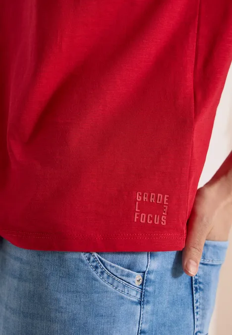 Rotes T-Shirt mit dezenter 'Garde Focus'-Stickerei am Saum und Jeanshosentasche, Hand lässig in der Tasche.