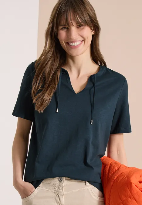 Frau in dunkelblauem Kurzarm-Kapuzenshirt und beiger Hose, mit orangefarbener Decke lässig am Arm.