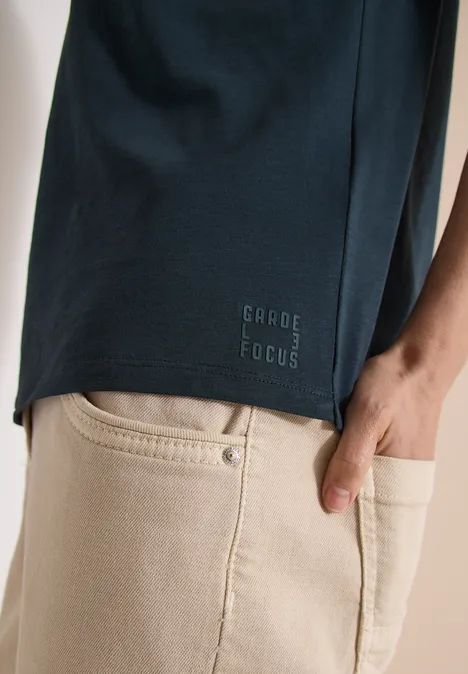 Ein dunkelblaues T-Shirt mit „GABE FOCUS“-Schriftzug am Saum, beige Jeans und eine Hand in der Gesäßtasche.