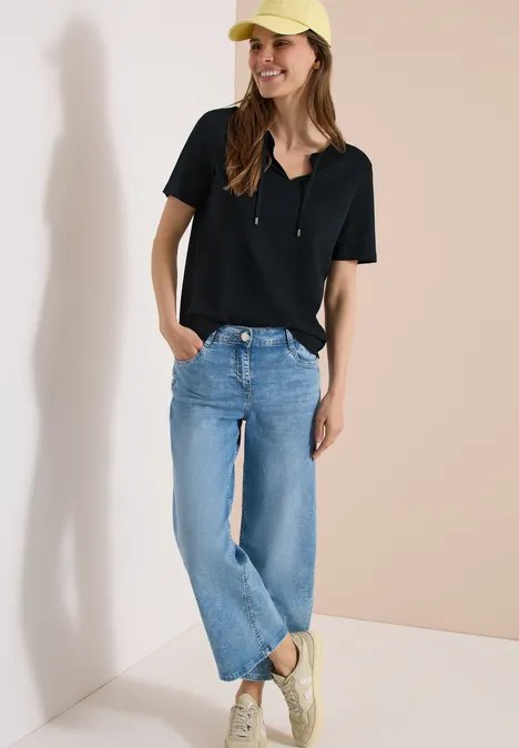 Stehende Frau in schwarzem Kurzarm-Top, hellen Jeans und beigen Slip-Ons vor hellem Hintergrund.