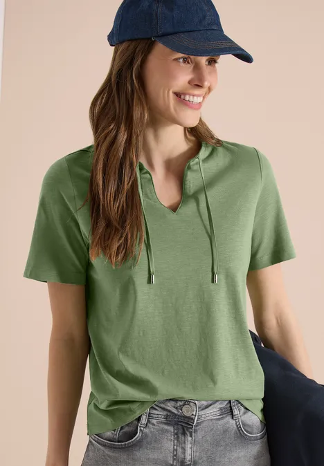 Person trägt olivgrünes Kapuzen-T-Shirt, Jeans und Schirmmütze vor hellem Hintergrund.