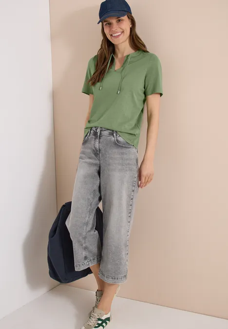 Eine Person trägt ein grünes Poloshirt, weite graue Jeans und eine blaue Kappe, hält eine Jacke in der Hand.