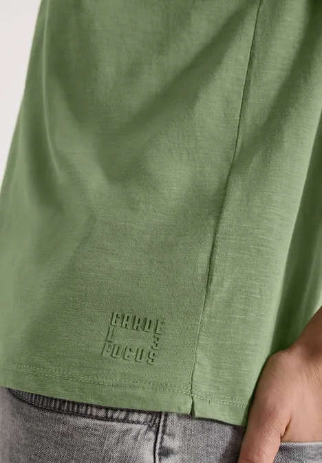 Grünes T-Shirt mit kleinem 'pearl focus'-Logo am Saum, Hand an der Hüfte.