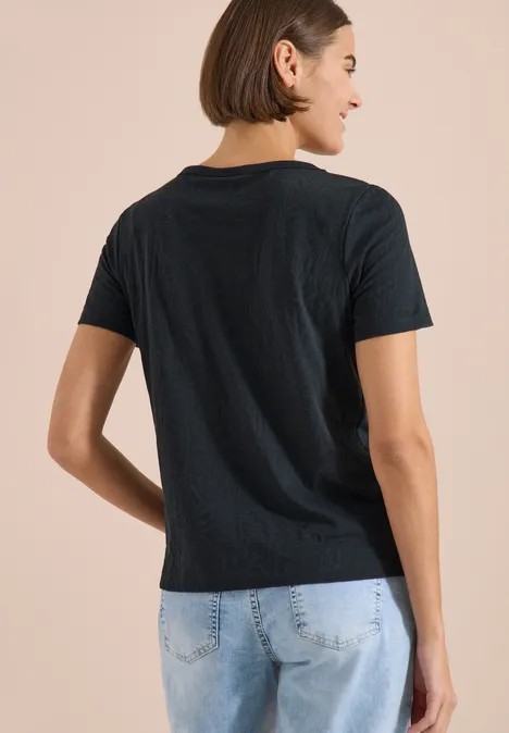 Shirt met korte mouwen in burnout-look palm breeze green Persoon van achteren in zwart t-shirt en lichte spijkerbroek tegen een beige achtergrond.