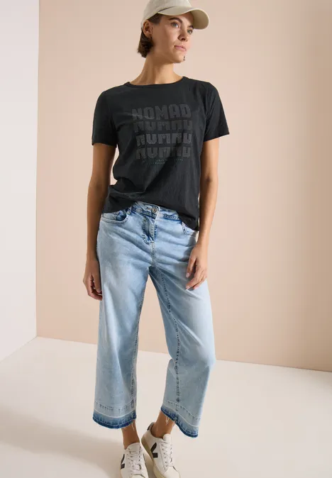 Shirt met korte mouwen in burnout-look palm breeze green Modieuze look: persoon in zwart T-shirt met print, lichte wijdvallende jeans, witte sneakers en pet.