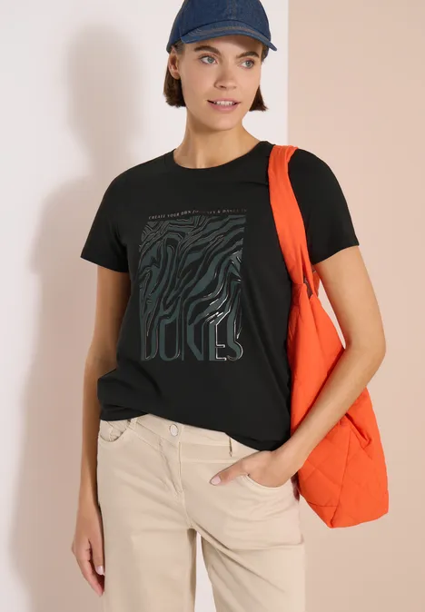 Person mit schwarzem T-Shirt und beigen Hosen trägt eine orangefarbene Tasche und eine Kappe.