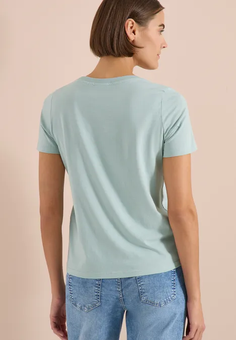 Frau von hinten in hellblauem T-Shirt und Jeans, schulterlanges braunes Haar.