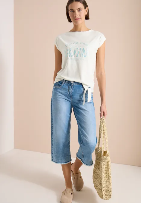 Frau mit weißem T-Shirt, weiten Culotte-Jeans, Basttasche und Espadrilles in heller Umgebung.
