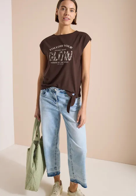 Frau in braunem T-Shirt, lockerer Jeans und grünem Stoffbeutel vor hellem Hintergrund.