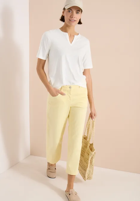 Shirt met gespleten hals in effen kleur vanilla white Vrouw in witte V-hals T-shirt, lichtgele broek en beige sneakers, met gevlochten strozak voor een lichte achtergrond.