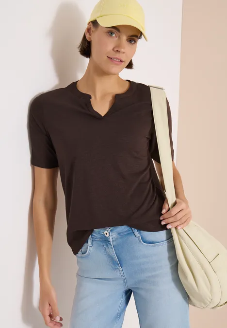T-Shirt mit Split Neck in Unifarbe brown cacao Frau in dunklem T-Shirt, hellblauen Jeans, hellbeigem Schulterbeutel und Cap vor zweifarbiger Wand