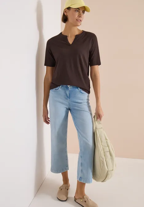 T-Shirt mit Split Neck in Unifarbe brown cacao Frau in schwarzem V-Shirt, hellen Jeans, beigen Slip-ons und großer Tasche vor neutralem Hintergrund.