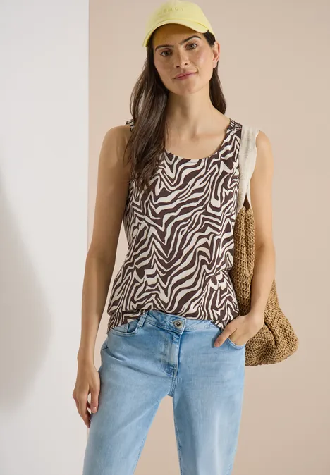 Top met ronde hals en plooidetail brown cacao Vrouw in een zebraprint top, lichtblauwe spijkerbroek en stro-tas voor een neutrale achtergrond.