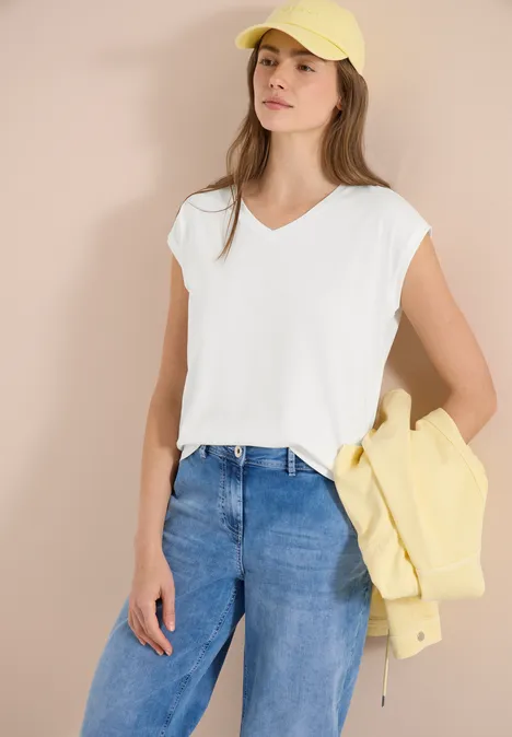 Frau trägt weiße V-Neck-Bluse, blaue Jeans, gelbe Jacke über dem Arm und gelben Baseballcap vor hellem Hintergrund.