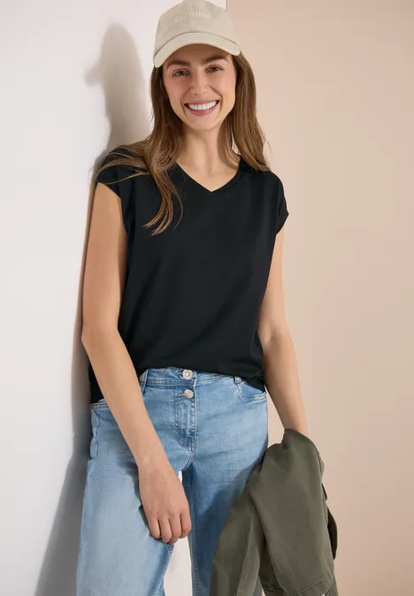 Chemise dropped shoulder avec col en V palm breeze green Frau trägt schwarzes V-Neck-T-Shirt mit kurzen Ärmeln, blaue Jeans, hält olivgrüne Jacke in der Hand.
