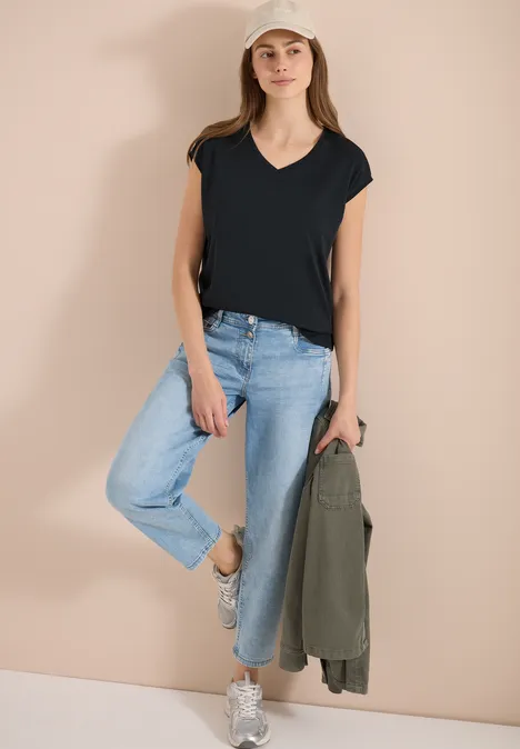 Chemise dropped shoulder avec col en V palm breeze green Frau lehnt an hellem Hintergrund, trägt schwarzes T-Shirt, blaue Jeans, weiße Sneakers und hält eine grüne Jacke.