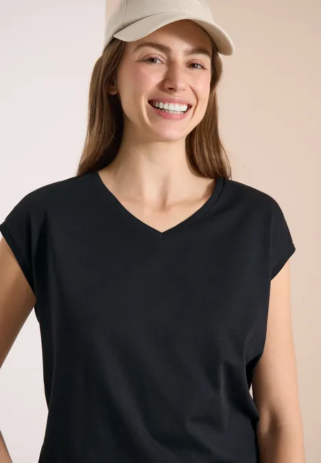 Chemise dropped shoulder avec col en V palm breeze green Frau mit schwarzem V-Neck-Shirt und hellbeiger Baseballkappe vor neutralem Hintergrund.