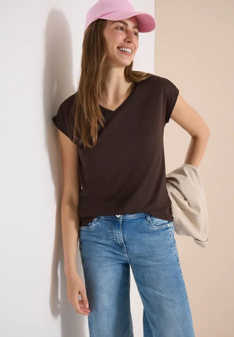 Frau trägt braunes T-Shirt, blaue Jeans und rosa Cap, lehnt lässig mit Jacke über dem Arm an heller Wand.