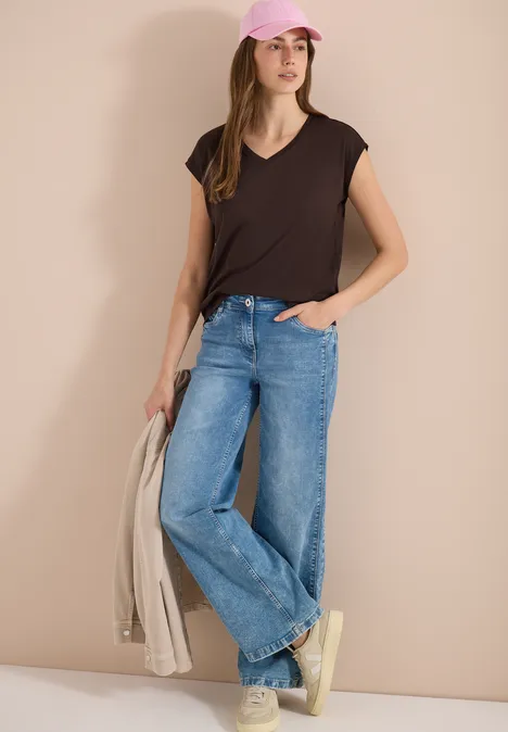 Frau in schwarzem T-Shirt, blauen Jeans und Sneakern hält eine beige Jacke.