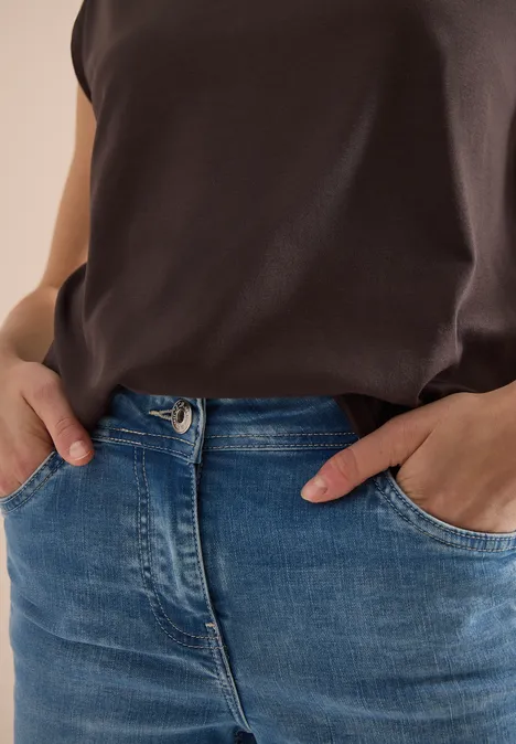 Eine Person trägt ein dunkelbraunes Oberteil und blaue Jeans, Hände in den Taschen.