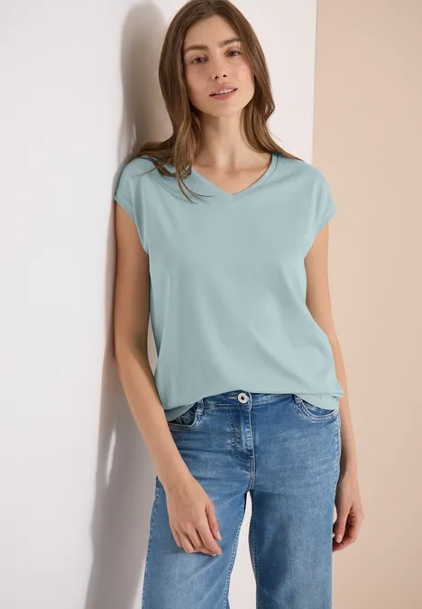 Shirt met overhangende schouders en V-hals crystal mint Een vrouw leunt tegen een muur en draagt een lichtblauw T-shirt met een spijkerbroek.
