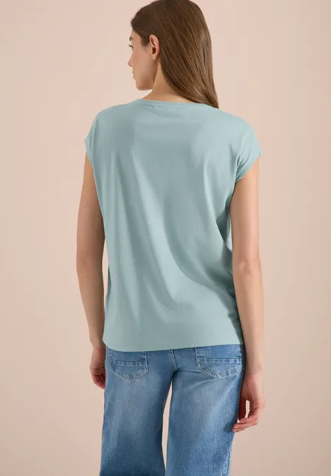 Shirt met overhangende schouders en V-hals crystal mint Vrouw vanachter in lichtblauw losvallend T-shirt en blauwe jeans tegen neutrale achtergrond.