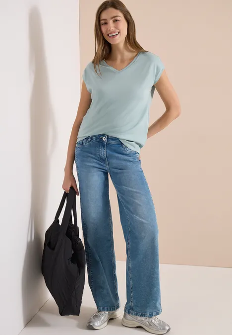 Shirt met overhangende schouders en V-hals crystal mint Een vrouw leunt tegen een muur, draagt een lichtblauw T-shirt, wijde jeans, witte sneakers en een zwarte tas.