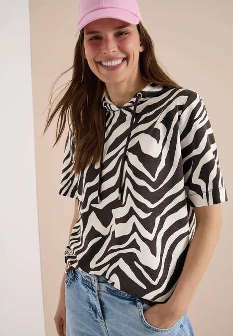 Vrouw voor lichte achtergrond draagt kort mouw capuchonshirt met zwart-witte zebraprint en blauwe spijkerbroek.