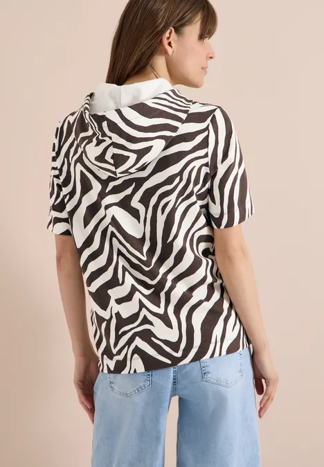 Vrouw van achteren in een kortmouwig zebraprintshirt met capuchon en lichtblauwe spijkerbroek tegen een neutrale achtergrond.