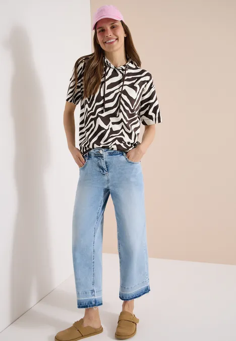 Vrouw staat voor neutrale achtergrond in zebraprintblouse, lichte wijde spijkerbroek en bruine sandalen.