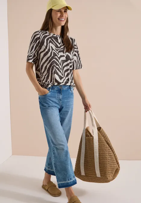 Frau trägt Zebramuster-Shirt, blaue Jeans, Pantoffeln und eine große geflochtene Strandtasche.